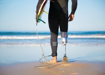 Unrecognizable disabled surfer going to water and carrying longboard. Cropped strong man wearing sportive swimsuit and holding surfboard. Physical disability, surfing and extreme sport concept