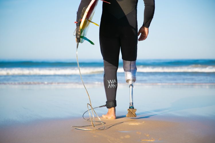 Unrecognizable disabled surfer going to water and carrying longboard. Cropped strong man wearing sportive swimsuit and holding surfboard. Physical disability, surfing and extreme sport concept